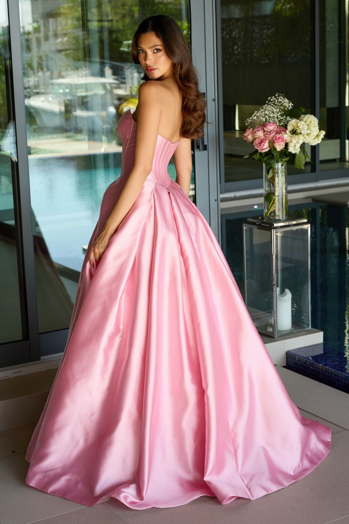 A woman wearing the Ava Presley 47223 strapless satin prom ball gown stands indoors by a glass door, with a vase of flowers and a pool visible in the background.